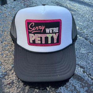 May include: A trucker hat with a white front panel, black brim, and mesh back. The front features a pink rectangle with the words "Sorry we're Petty" in a playful font. The hat is designed for casual wear.