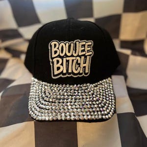 May include: Black baseball cap with the text "BOUJEE BITCH" in white and gold lettering. The brim is fully encrusted with sparkling rhinestones. The hat is set against a black and white checkered background.