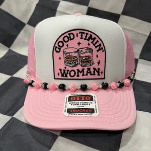 May include: Pink trucker hat with a white front panel featuring the text "GOOD TIMIN WOMAN" and an illustration of two glasses. The hat has a pink mesh back and a beaded brim with pink and black beads.