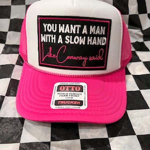 May include: A pink trucker hat with a white front panel featuring the text "YOU WANT A MAN WITH A SLOW HAND Like Conway said" in black and pink. The hat has a pink brim and mesh back, with a snap closure.