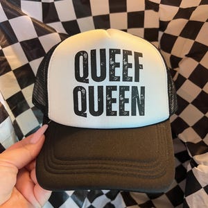 May include: A black and white trucker hat featuring a white foam front panel with the black text QUEEF QUEEN printed in a bold, distressed sans-serif font. The hat includes a black brim and black mesh side and back panels for ventilation.