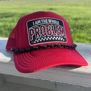 May include: Red trucker hat with the text "I AM THE WHOLE PROBLEM" in a black and white design. The hat has a black chain detail across the brim and a mesh back.