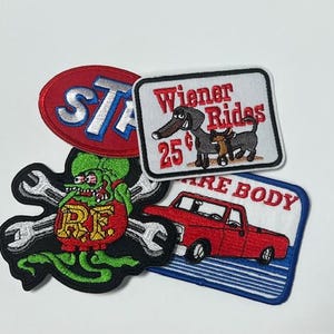 May include: A collection of embroidered patches with various designs. One patch features a red oval with "STA" in white and blue. Another shows a cartoon dachshund with the text "Wiener Rides 25¢". Other patches include a monster and a red truck.