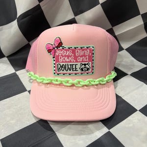 May include: Pink trucker hat with a mesh back and a curved brim. The hat features a patch with the text "Jesus, Bling, Bows, and Boujee" and a raccoon graphic. A green chain decorates the brim.