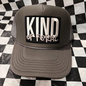 May include: A gray trucker hat with a mesh back and a curved brim. The front features a rectangular patch with the words "KIND of FERAL" in white and gray lettering. The hat is set against a black and white checkered background.
