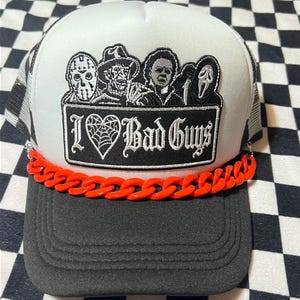 May include: A white and black trucker hat featuring a rectangular patch with illustrations of four horror movie characters above the text I Heart Bad Guys, where the heart is a spiderweb. A bright orange plastic chain decorates the brim base.