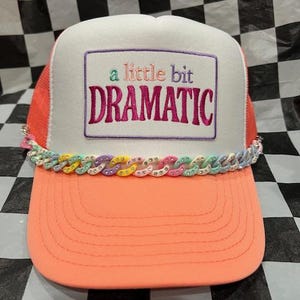 May include: A trucker hat with a white front panel, a coral-colored brim, and a mesh back. The hat features the embroidered phrase "a little bit DRAMATIC" and a colorful chain detail across the brim.