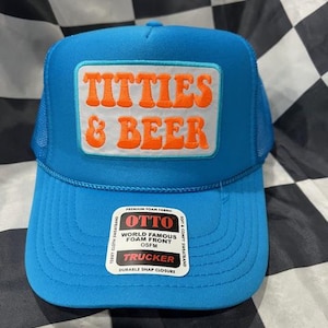May include: A blue trucker hat with a white patch featuring the words "TITTIES & BEER" in orange. The hat has a foam front and a mesh back. The hat also has a tag that says "OTTO" and "TRUCKER".