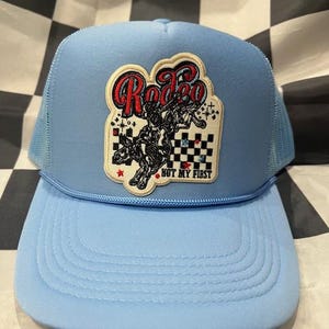 May include: Light blue trucker hat with a curved brim and mesh back. The front features a patch with the word "Rodeo" in red script and a black and white graphic of a rodeo rider. The text "NOT MY FIRST" is also visible.