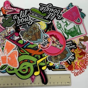 May include: A collection of colorful embroidered patches with various designs, including a bow, musical notes, and text. The patches feature a variety of colors, including pink, green, orange, and yellow. A ruler is at the bottom, measuring in inches.