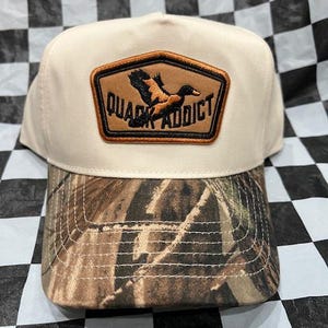 May include: A beige baseball cap with a camouflage brim. The cap features a brown patch with the words "QUACK ADDICT" and a flying duck graphic.