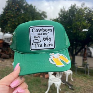 May include: Green trucker hat with a white patch that reads "Cowboys and beer that's why I'm here." The hat also features two beer mugs and a silver zipper.