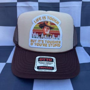 May include: A brown and tan trucker hat with a patch featuring a portrait of a man in a cowboy hat and the text "LIFE IS TOUGH BUT IT'S TOUGHER IF YOU'RE STUPID." The hat has a foam front and a snap closure.