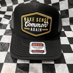 May include: Black trucker hat with a mesh back and a flat brim. The front features a gold and white patch that reads "MAKE SENSE COMMON AGAIN". The hat includes a "OTTO" tag.
