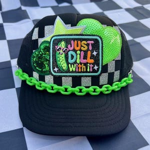 May include: Black trucker hat with a checkered pattern and a curved bill. The hat features a patch with a cartoon pickle wearing sunglasses and the text "JUST DILL WITH IT." Embellished with green chain, stars, and hearts.