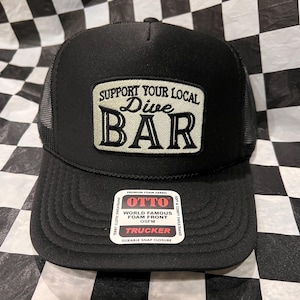 May include: Black trucker hat with a curved brim and mesh back. The front patch reads "SUPPORT YOUR LOCAL Dive BAR" in a vintage style. The hat includes a "OTTO" tag with "TRUCKER" printed on it.