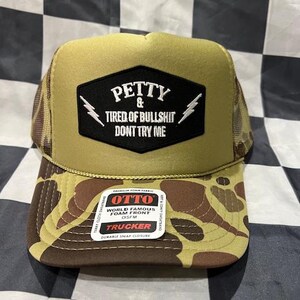 May include: A trucker hat with a camouflage pattern on the brim and a tan front panel. The front features a black patch with white text that reads "PETTY & TIRED OF BULLSHIT DONT TRY ME" and lightning bolt graphics. The hat has a mesh back.