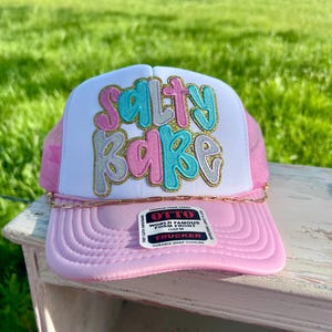 May include: A pink and white trucker hat with the text "Salty Babe" in colorful, glittery letters. The hat has a pink brim and mesh backing, with a gold chain detail. The hat is sitting on a white surface.