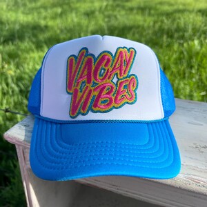 May include: A blue and white trucker hat with the words "VACAY VIBES" in pink and yellow embroidery. The hat has a blue brim and mesh back, perfect for sunny days and outdoor activities.