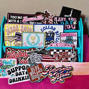 May include: A display of embroidered patches with various designs and text, including "Jesus Saves," "American Made," and "Have the Day You." The patches are in a turquoise organizer on a pink surface.