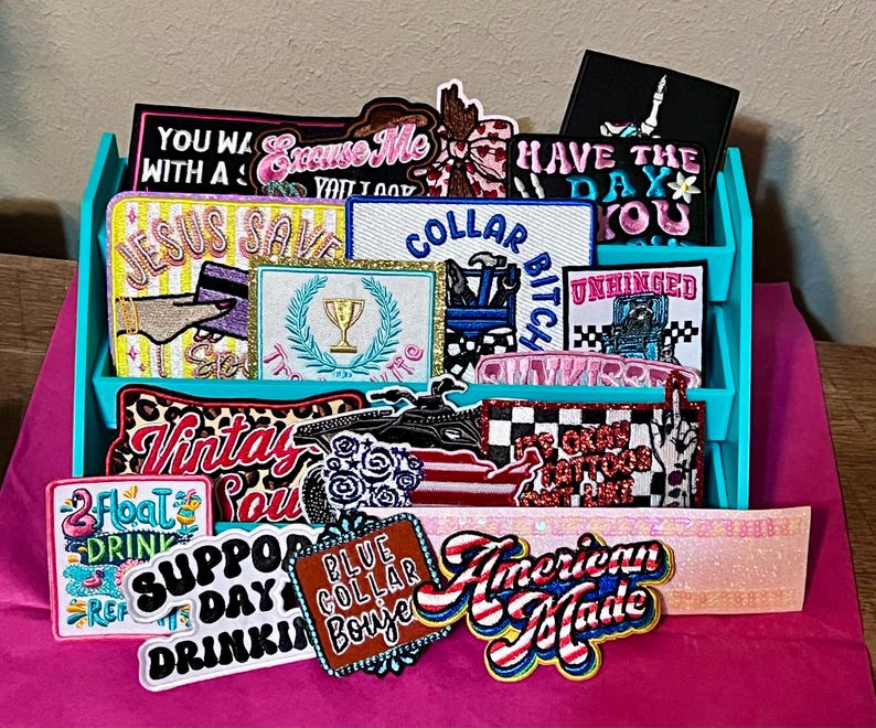 Pode incluir: Uma exibi&ccedil;&atilde;o de patches bordados com v&aacute;rios designs e textos, incluindo "Jesus Saves" e "American Made". Os patches est&atilde;o em um organizador turquesa sobre uma superf&iacute;cie rosa.