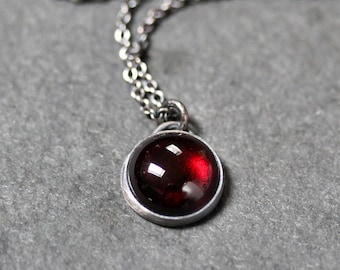 May include: A silver necklace featuring a round, deep red gemstone pendant. The pendant is set in a silver bezel and suspended from a delicate silver chain. The gemstone has a polished surface, and the chain is visible against a grey background.
