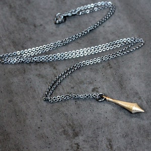 Puede incluir: Un collar de cadena de plata con un colgante puntiagudo de color dorado. El colgante tiene forma de triángulo puntiagudo y alargado.