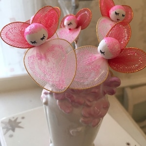 May include: Four pink and white fairy figurines with closed eyes and gold wire wings in a white vase with pink floral details.