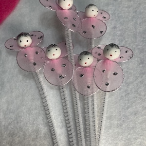 May include: A collection of handmade fairy ornaments. Each fairy has a white head with black eyes and a red mouth, attached to pink, glitter-edged wings. The wings are connected to a silver pipe cleaner stem.
