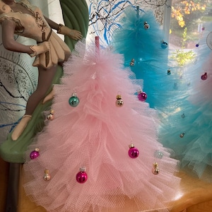 May include: Pink and blue tulle Christmas trees decorated with small ornaments. The pink tree has pink and gold ornaments, while the blue tree has silver and blue ornaments. A decorative statue is in the background.