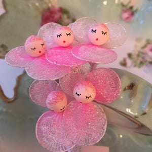 May include: A cluster of pink and white glitter flower-shaped ornaments with small pink faces and closed eyes. The ornaments are attached to thin wires.