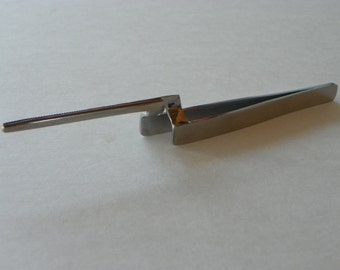 May include: A pair of silver-toned tweezers with a long, thin arm extending from the handle. The tweezers are designed for precision work.