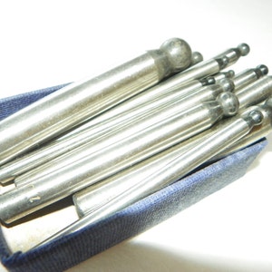 May include: A set of metal tools with rounded tips, arranged in a blue velvet-lined box. The tools are silver in color and have varying lengths.