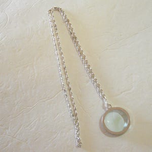 May include: A silver-toned necklace with a circular pendant. The pendant has a clear, light-green center and a silver frame. The chain is a twisted rope design.