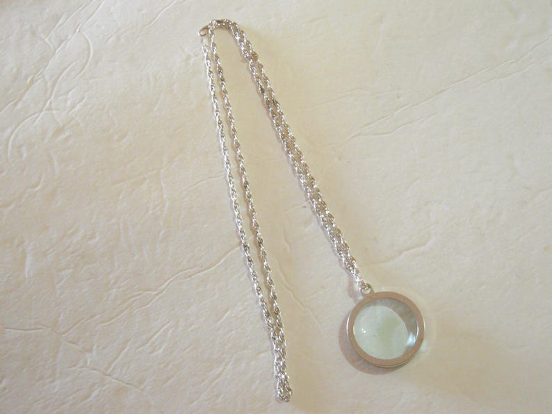 May include: A silver-toned necklace with a circular pendant. The pendant has a clear, light-green center and a silver frame. The chain is a twisted rope design.