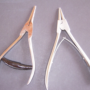May include: Two metal pliers, one with a black handle and the other with a silver handle. The pliers have pointed tips and curved handles. The tools are likely used for gripping, bending, or cutting materials. The image is taken on a gray surface.