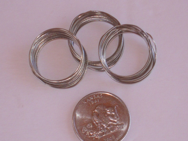 May include: Three spools of silver-colored wire, each wound into a circle, are arranged on a white surface. A silver US quarter is placed below the spools for scale.
