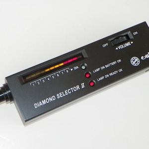 May include: A black Diamond Selector II tool with a pen-like tip. The device has a digital display with a color-coded scale and the text "DIA". It features a volume control and indicator lights for battery and readiness.