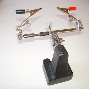May include: A silver metal workbench with two adjustable clamps. One clamp has a black handle and the other has a red handle. The workbench has a black cast iron base.