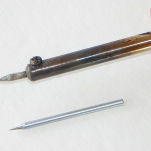 May include: A soldering iron with a red handle and a metallic shaft featuring a pointed tip secured by a small screw, accompanied by a separate, clean, silver-colored replacement soldering tip lying parallel below the main tool.
