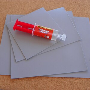 K&ouml;nnte beinhalten: Vier graue quadratische Bl&auml;tter aus Material mit einer durchsichtigen Epoxid-Klebstofftube mit der Aufschrift "Superseal Epoxy Clear Adhesive" darauf. Die Tube hat ein rotes und wei&szlig;es Etikett mit einem Uhrsymbol.