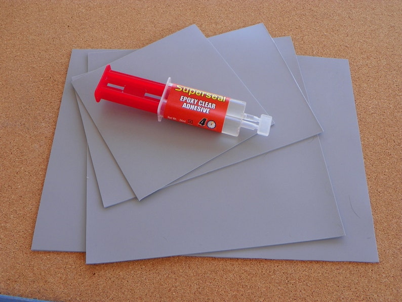 K&ouml;nnte beinhalten: Vier graue quadratische Bl&auml;tter aus Material mit einer durchsichtigen Epoxid-Klebstofftube mit der Aufschrift "Superseal Epoxy Clear Adhesive" darauf. Die Tube hat ein rotes und wei&szlig;es Etikett mit einem Uhrsymbol.