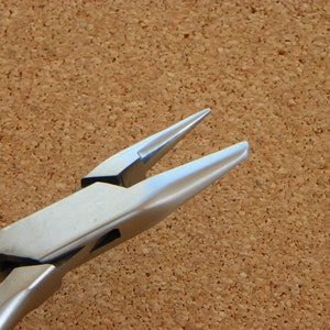 May include: Close-up of a pair of silver-toned pliers with a pointed tip. The pliers are on a brown cork surface.