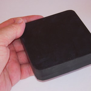 May include: A black, square object with rounded corners is held in a hand. The object appears to be made of a solid, matte material. The image is taken against a plain white background.