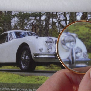 May include: A white vintage car is the main subject of the image, captured in a photograph. The car is viewed through a magnifying glass with a gold rim, held by a person. The car is parked on a grassy area with trees in the background.