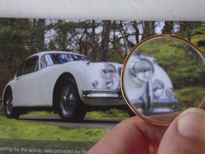 May include: A white vintage car is the main subject of the image, captured in a photograph. The car is viewed through a magnifying glass with a gold rim, held by a person. The car is parked on a grassy area with trees in the background.
