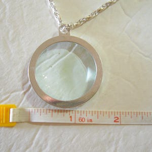May include: A silver-toned pendant necklace with a clear, circular locket containing a light-colored object. The necklace chain is a twisted rope design. A measuring tape shows the pendant is approximately 2 inches in diameter.