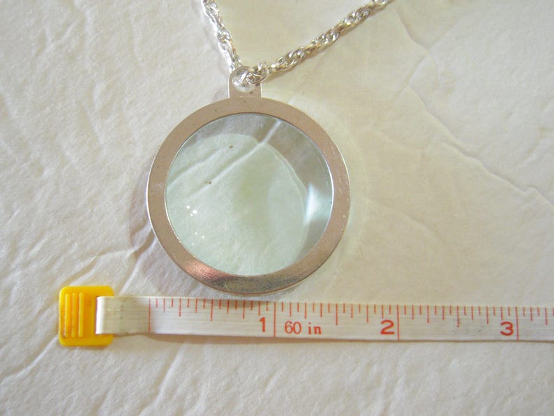 May include: A silver-toned pendant necklace with a clear, circular locket containing a light-colored object. The necklace chain is a twisted rope design. A measuring tape shows the pendant is approximately 2 inches in diameter.