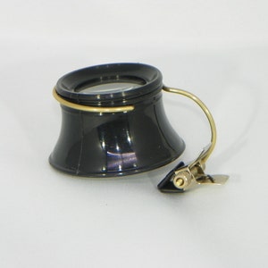 May include: A black magnifying glass with a gold-colored rim and a clip for attaching to a surface. The magnifying glass is designed for hands-free use.