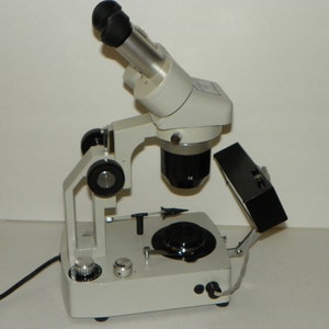 May include: A white and black microscope with a black light source. The microscope has a black eyepiece and a black knob on the side. The light source is attached to the microscope with a black arm.